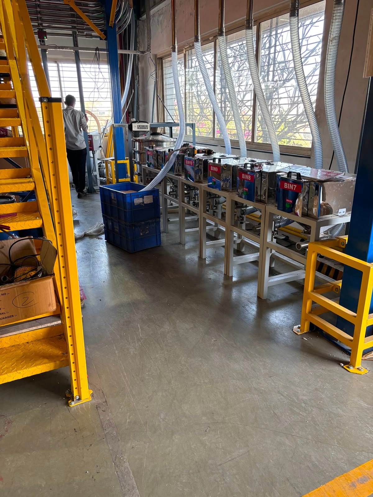 Row of stainless steel day bins connected to flexible feed hoses in a production area